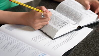Persona consultando un diccionario con un lápiz en su mano y un libro en la mesa