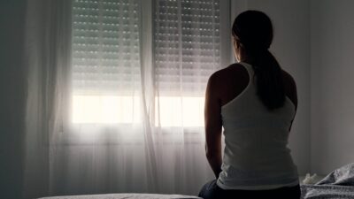 Mujer sentada en cama observando cabizbaja una ventana a medio abrir