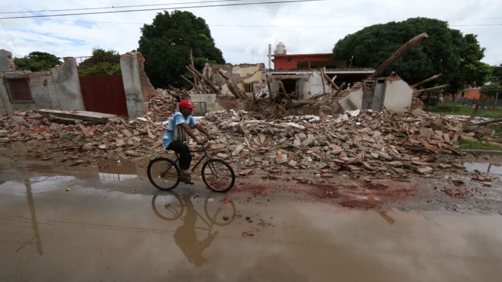 Hombre en bicicleta pasea por una calle afectada por un sismo. En el fondo se ve una barda derrumbada