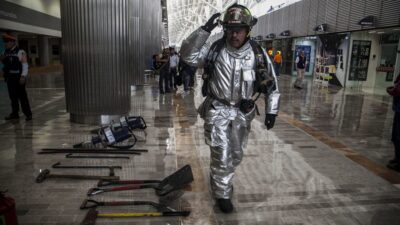 Bombero con traje plateado y herramientas diversas a su lado, en el suelo, en el Aeropuerto de la CDMX