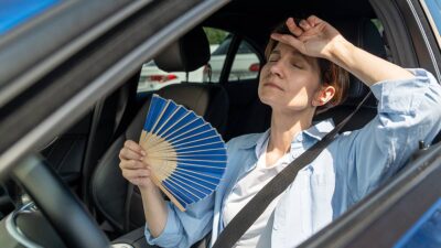 Mujer conduciendo su auto bajo el sol, con la ventanilla abierta y con un abanico para el calor